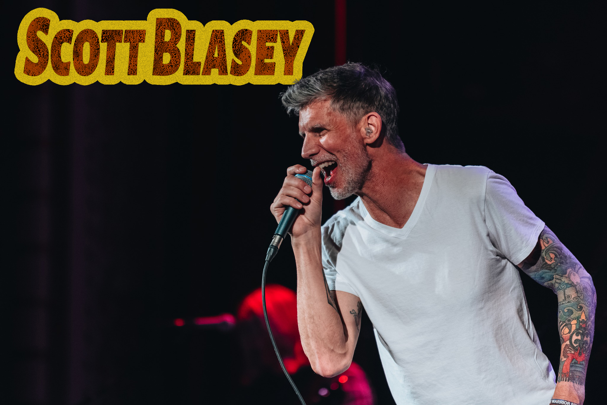Man with tattoos sings into microphone on stage; Scott Blasey text appears in the corner.