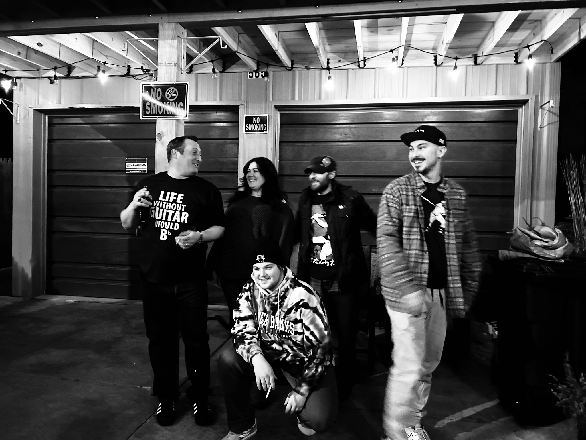 Five people pose and laugh together outside by a garage under No Smoking signs at night.